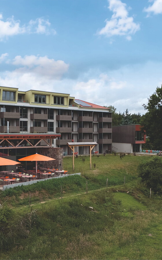Weitwinkelaufnahme des grünen Geländes der Seeziege Thomsdorf, das direkt an einen klaren See grenzt, mit Bäumen und einem Teil der Anlage im Hintergrund.