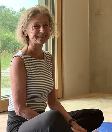 Eine lächelnde Frau mit kurzen blonden Haaren sitzt im gestreiften Oberteil und dunkler Hose barfuß im Schneidersitz auf einer blauen Yogamatte.